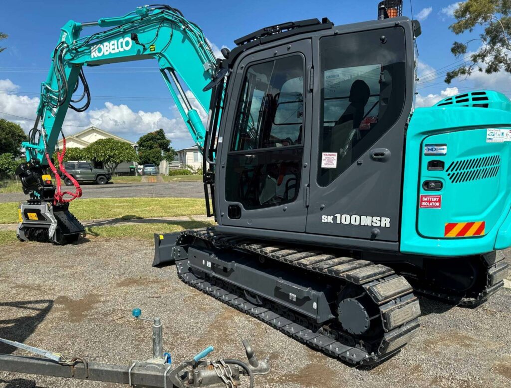 A Kobelco SK100MSR excavator with a teal and black body is positioned on a gravel surface. The machinery has a large arm extending upwards. Buildings and trees can be seen in the background under a partly cloudy sky.