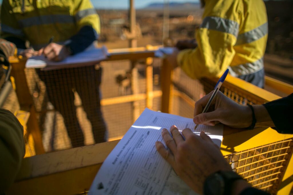 Workers in high-visibility clothing fill out forms on clipboards, standing around a metal railing outdoors. The background appears to be an industrial or construction site with blurred structures and earth tones visible.