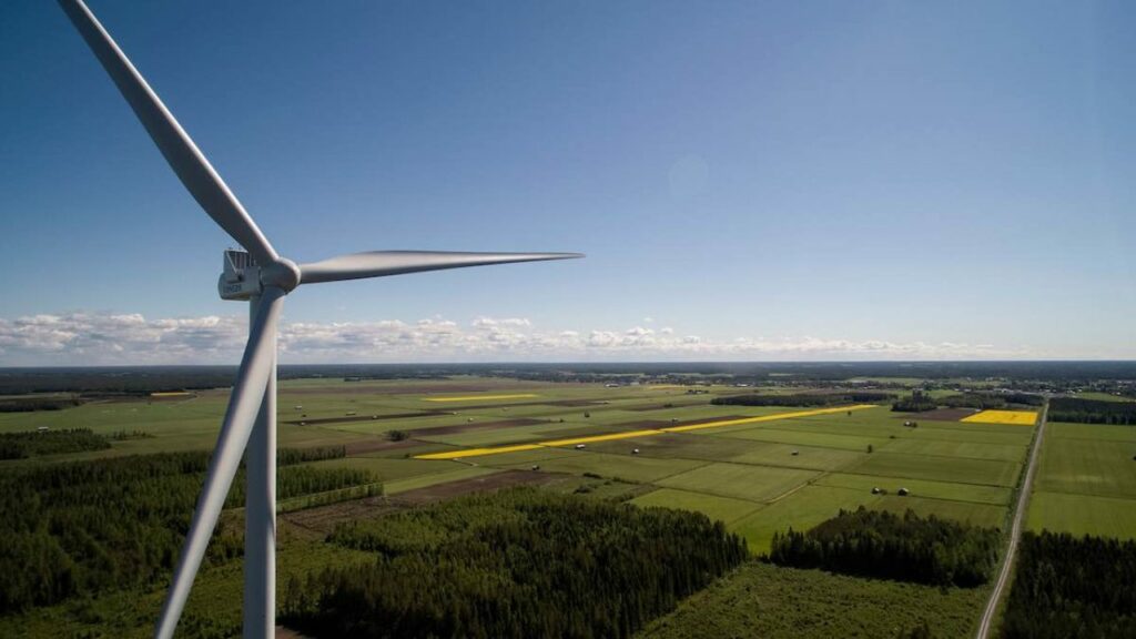 Aerial view of a wind turbine standing tall over expansive green fields and wooded areas under a clear blue sky. The landscape features patches of yellow, possibly rapeseed or similar crops, and several distant farm buildings.