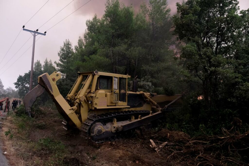 A yellow bulldozer is clearing vegetation near a forest fire. Thick smoke billows from the trees in the background. Power lines run alongside a nearby road, with a few people visible in the distance, observing the scene. Dense green foliage surrounds the area.