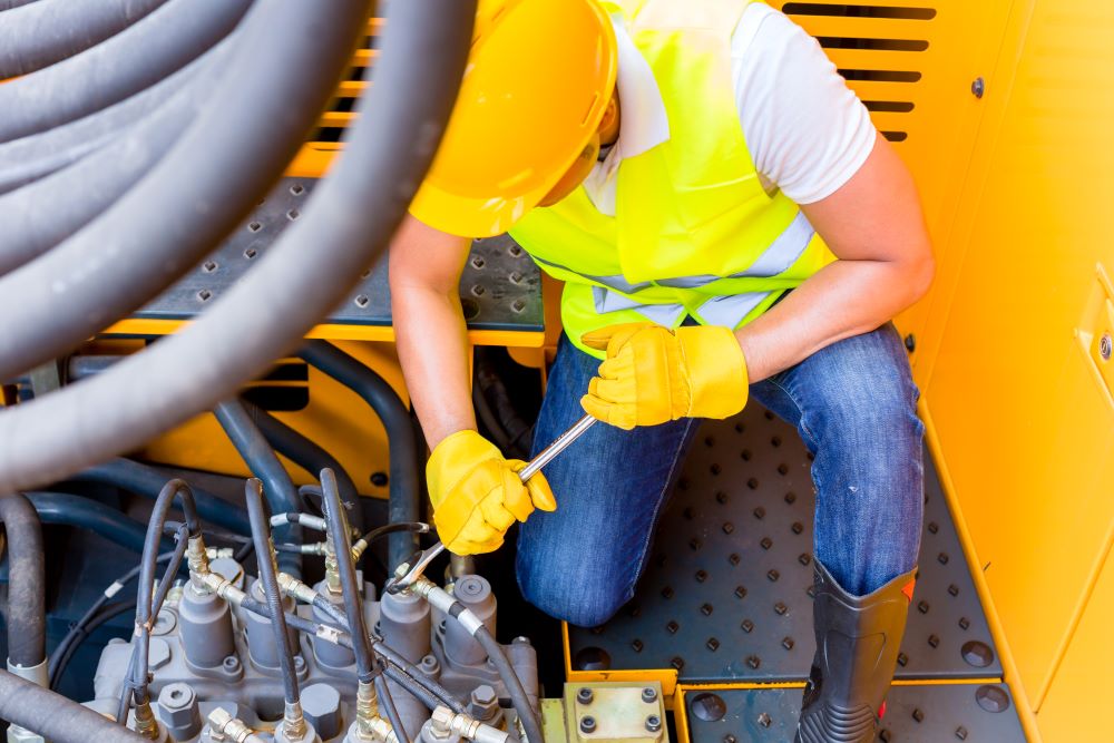 A worker in a high-visibility vest and yellow hard hat, specializing in truck hire services, uses a wrench to adjust machinery in an industrial setting. Large black cables and various mechanical components surround the worker, who is kneeling and focused on the task.