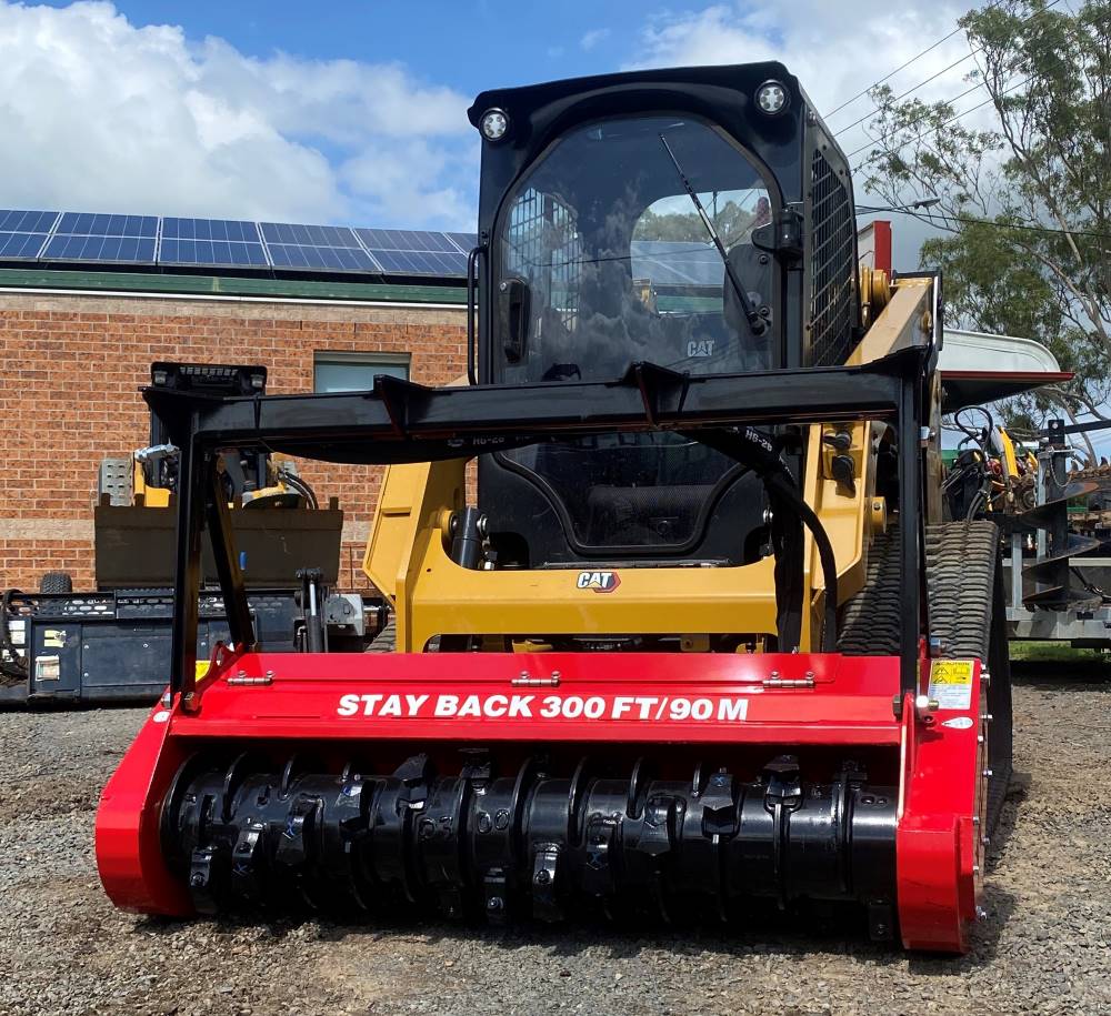 A yellow CAT skid steer loader equipped with a red forestry mulcher attachment is parked on a gravel area. The mulcher has a warning text that reads "STAY BACK 300 FT/90 M." A brick building with solar panels on the roof is in the background, alongside a sign for Truck Hire services.