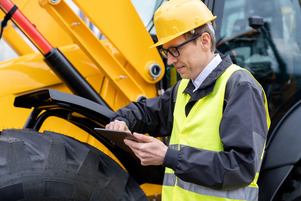 A construction worker in a yellow hard hat and reflective vest stands beside heavy machinery, focusing on a tablet. The person appears to be inspecting or logging information about the equipment, possibly coordinating truck hire services.
