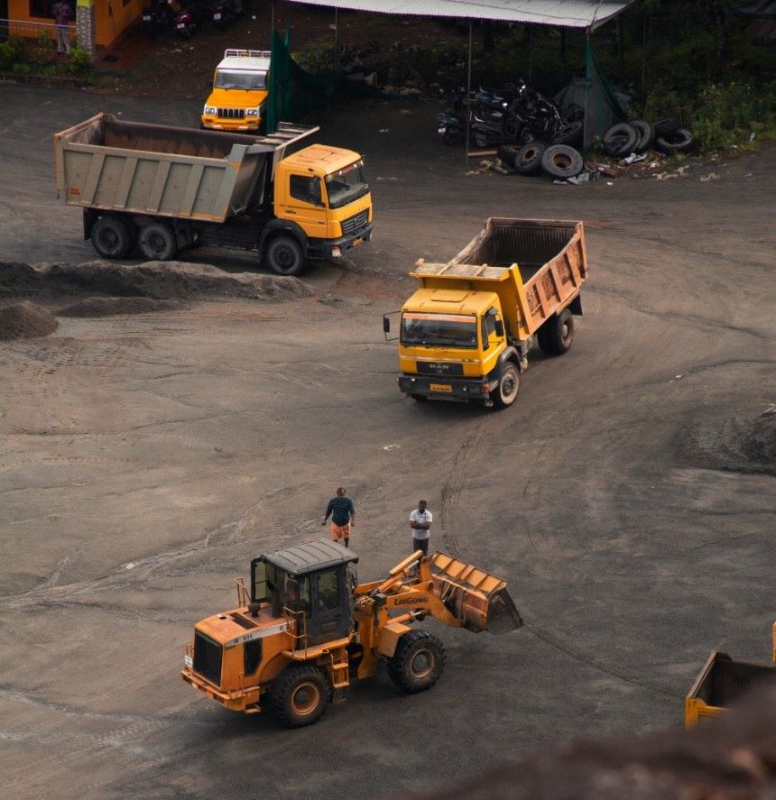 Aerial view of a construction site with two yellow dump trucks available for truck hire and one front loader. Two workers are standing near the front loader, which has its bucket partially raised. The ground appears covered with dark gravel or dirt. Several motorcycles are parked in the background.